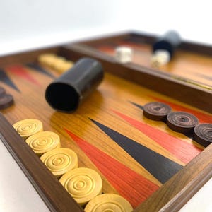 May include: A wooden backgammon board with a warm, reddish-brown finish. The game pieces include light tan and dark brown checkers, dice, and a black doubling cube. The board features red and black triangular points.