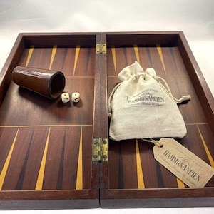 Puede incluir: Un juego de backgammon de madera con acabado marrón oscuro e incrustaciones amarillas. Incluye un cubilete marrón, dos dados y una bolsa con la inscripción "GAMMONANCIEN". También hay una etiqueta con el mismo texto.