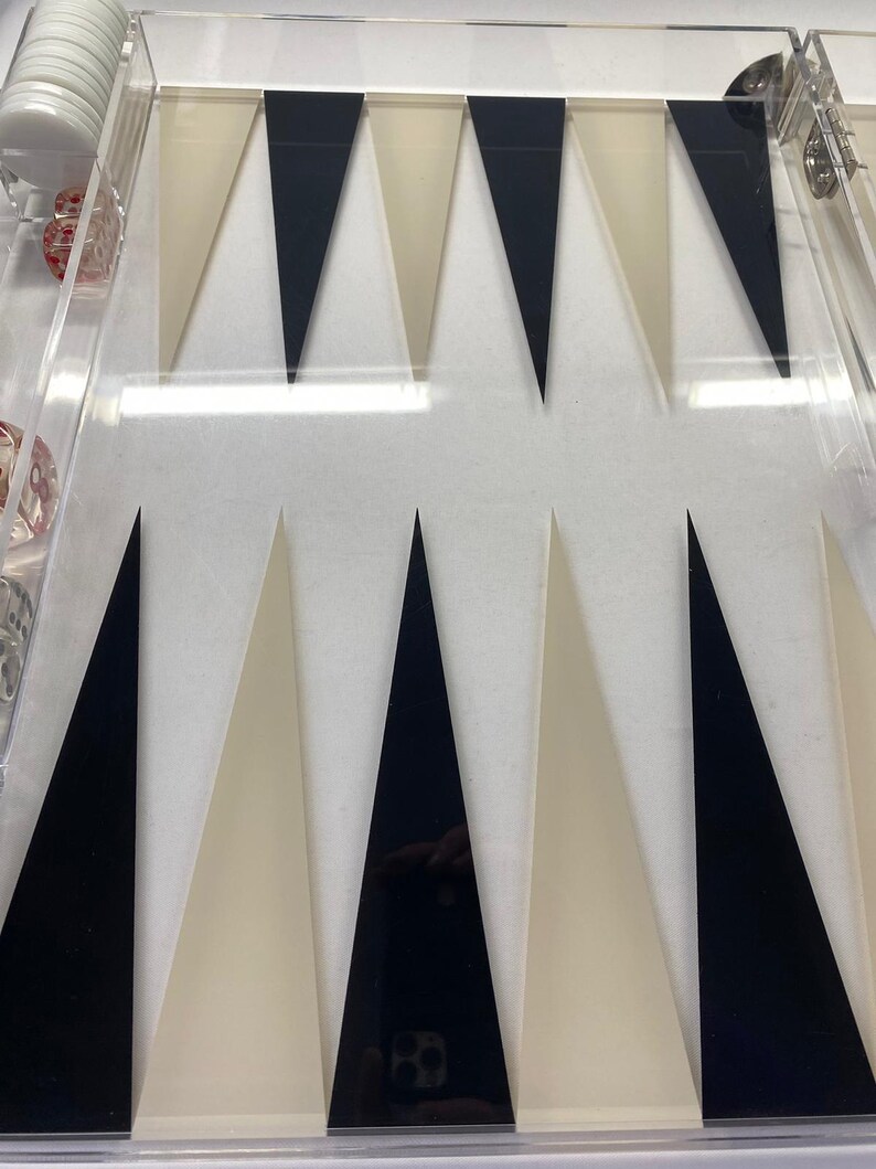 Pode incluir: Um tabuleiro de backgammon em acr&iacute;lico transparente com pontas triangulares pretas e creme. O tabuleiro inclui pe&ccedil;as brancas, dados vermelhos e um copo para dados. O jogo &eacute; projetado para dois jogadores.