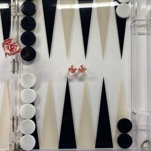 Pode incluir: Um conjunto de gam&atilde;o em acr&iacute;lico transparente com pe&ccedil;as pretas e brancas. O tabuleiro apresenta tri&acirc;ngulos pretos, brancos e creme alternados. Dois dados vermelhos com pontos brancos e um dado grande est&atilde;o inclu&iacute;dos.