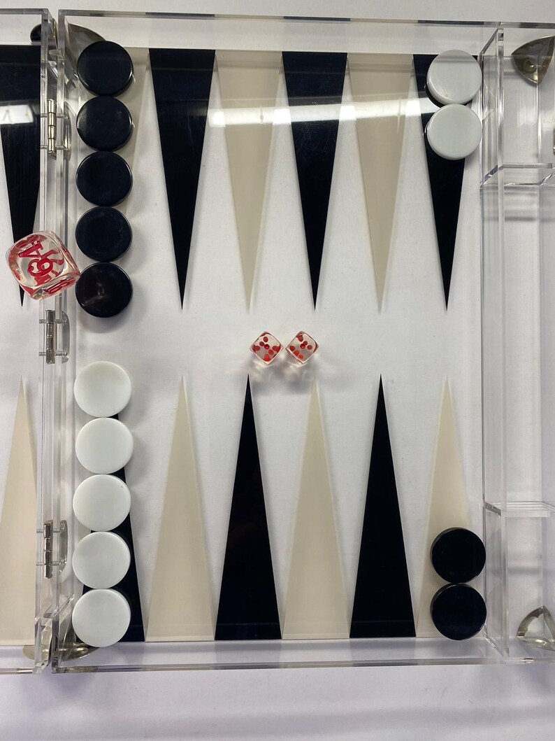 Pode incluir: Um conjunto de gam&atilde;o em acr&iacute;lico transparente com pe&ccedil;as pretas e brancas. O tabuleiro apresenta tri&acirc;ngulos pretos, brancos e creme alternados. Dois dados vermelhos com pontos brancos e um dado grande est&atilde;o inclu&iacute;dos.