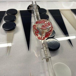 Pode incluir: Um tabuleiro de gam&atilde;o com tri&acirc;ngulos pretos e brancos, pe&ccedil;as pretas e um dado transparente com n&uacute;meros vermelhos. O tabuleiro &eacute; feito de acr&iacute;lico transparente e tem uma dobradi&ccedil;a de metal.