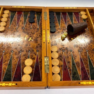Puede incluir: Un juego de backgammon de madera con una superficie de juego con incrustaciones. El juego abierto revela fichas negras y beige, dados y un cubilete. El tablero presenta una rica paleta de colores con acentos rojos, verdes y marrones.