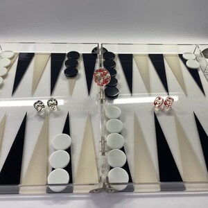 Pode incluir: Um conjunto de gam&atilde;o em acr&iacute;lico transparente com pontas triangulares pretas, brancas e bege. O tabuleiro apresenta pe&ccedil;as pretas e brancas e tr&ecirc;s dados. Os dados s&atilde;o vermelhos e transparentes.