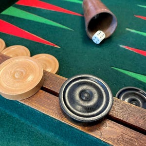 May include: Backgammon game in progress, featuring a green playing surface with red and green triangles. Wooden game pieces in black and tan are visible, along with a brown dice cup and a white die showing the number 6.