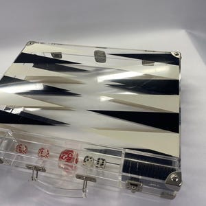 Pode incluir: Conjunto de backgammon em acr&iacute;lico transparente com um design geom&eacute;trico em preto e branco. O conjunto inclui dados vermelhos e brancos e uma caixa transparente com uma al&ccedil;a e fechos de metal. Um jogo de tabuleiro cl&aacute;ssico.