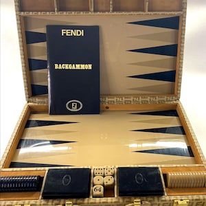 Puede incluir: Juego de backgammon Fendi abierto con un patrón de monograma beige y marrón. El juego incluye un folleto que dice "FENDI BACKGAMMON", dados y fichas de juego negras y beige. El estuche tiene un asa y cierres dorados.
