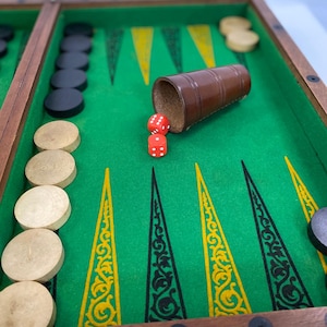 May include: A backgammon board with a green playing surface, wooden frame, and decorative yellow and black triangles. The game includes black and tan game pieces, two red dice, and a brown dice cup.