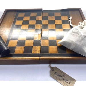 May include: A wooden backgammon board featuring a black and natural wood checkered pattern. The set includes a black cylindrical dice cup, a beige drawstring fabric pouch with the text GAMMONANCIEN, and a cardboard tag labeled GAMMONANCIEN.