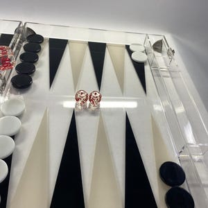 Pode incluir: Um conjunto de backgammon em acr&iacute;lico transparente com pontas triangulares pretas, brancas e bege. O conjunto inclui pe&ccedil;as pretas e brancas e dois dados vermelhos com pontos brancos. O tabuleiro &eacute; retangular e tem uma borda transparente.