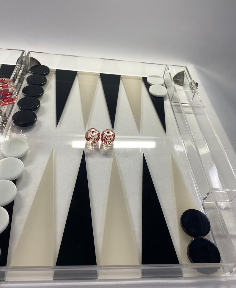 Pode incluir: Um conjunto de backgammon em acr&iacute;lico transparente com pontas triangulares pretas, brancas e bege. O conjunto inclui pe&ccedil;as pretas e brancas e dois dados vermelhos com pontos brancos. O tabuleiro &eacute; retangular e tem uma borda transparente.
