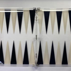 Pode incluir: Um conjunto de gam&atilde;o em acr&iacute;lico transparente com uma superf&iacute;cie de jogo branca. O tabuleiro apresenta tri&acirc;ngulos pretos e creme alternados. Dados, pe&ccedil;as e outras pe&ccedil;as do jogo s&atilde;o vis&iacute;veis nos compartimentos transparentes.