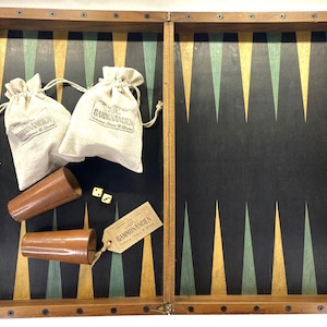 Könnte beinhalten: Ein Backgammon-Set aus Holz mit schwarzer Spielfläche, grünen und gelben Dreiecken. Enthält zwei Würfelbecher aus Leder, zwei Kordelzugbeutel, Würfel und ein Etikett. Das Set befindet sich in einem Holzrahmen.