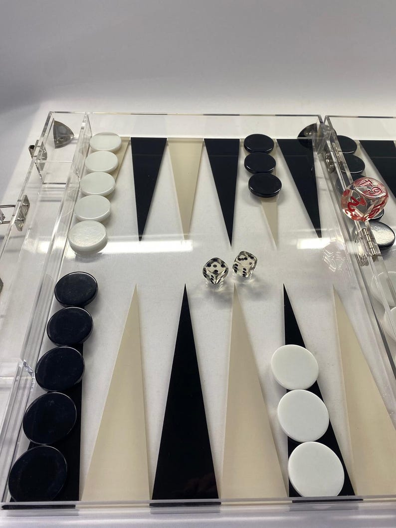 Pode incluir: Um conjunto de gam&atilde;o em acr&iacute;lico transparente com pontas triangulares pretas, brancas e bege. O conjunto inclui pe&ccedil;as pretas e brancas e dois dados. O tabuleiro &eacute; transparente.