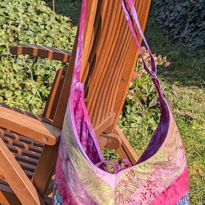 Könnte beinhalten: Handgefertigte Schultertasche mit Patchwork-Design in Pink-, Lila- und Grüntönen. Die Tasche hängt an einem Holzstuhl im Freien und zeigt verschiedene Stoffmuster.