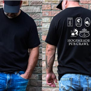 May include: Black t-shirt with a white graphic on the back. The graphic features the names and logos of five pubs: The Leaky Cauldron, The Three Broomsticks, Hogsmeade, The Green Dragon, and Hog's Head. The text "Hogsmeade Pub Crawl" is printed below the logos.