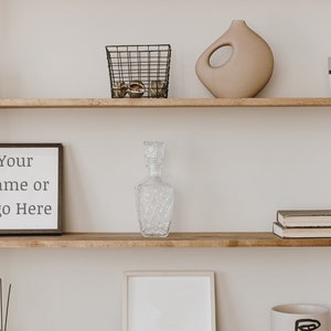 Peut inclure: Une étagère en bois avec un cadre noir contenant l'inscription "Your Name or Logo Here", une carafe en verre transparent, un vase en céramique marron, un panier en fil de fer avec des objets ronds, une bougie marron, une tasse blanche et deux livres.