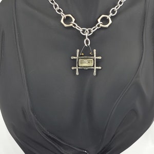 May include: A silver chain necklace with a hexagonal link pattern. The necklace features a pendant with a small, rectangular frame and a glass window with the text "1013C, 71" inside. The pendant is attached to the chain with a silver loop.
