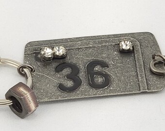 Pode incluir: Um chaveiro prateado com uma etiqueta de metal que diz "36" e tem três strass. O chaveiro também tem um charme de chave prateado e um charme de porca prateado.