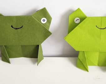 May include: Two green origami frogs with black eyes and a black smile on a white surface.