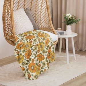 May include: A woven wicker hanging chair with a white throw pillow and a floral patterned throw blanket in shades of orange, yellow, and green. The chair is in a room with a white side table and a vase of white flowers.