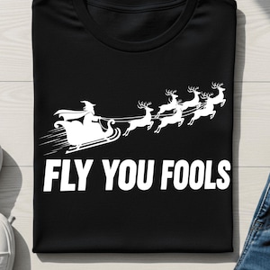 May include: Black t-shirt with a white graphic of a witch in a sleigh pulled by reindeer, with the text "FLY YOU FOOLS". The shirt is folded, with white sneakers and denim jeans visible.