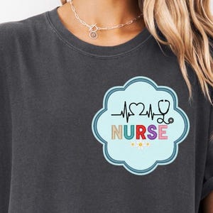 May include: Dark gray t-shirt with a colorful nurse-themed design. The design includes a heartbeat line, a stethoscope, and the word "NURSE" in a playful font. The shirt has rolled-up sleeves and is paired with a denim jacket. The bottom of the image reads "Digital Download PNG_SVG".