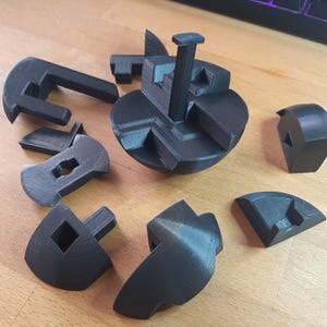 May include: A black 3D printed puzzle with multiple pieces. The puzzle is disassembled and laid out on a wooden surface. The pieces are various shapes and sizes, and some have holes or slots.