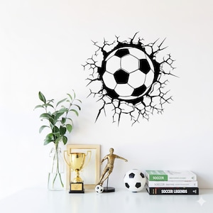 Może przedstawiać: Grafika czarno-białej piłki nożnej, która wydaje się przebijać przez ścianę, otoczona efektem pęknięcia. Obraz zawiera również złote trofeum, figurkę piłkarza i piłkę nożną.