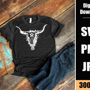 May include: A dark gray t-shirt with a white graphic of a stylized bull skull with a star in the center. The t-shirt is laying on a wooden surface with a pair of brown cowboy boots and blue jeans.