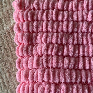 May include: Close-up of a pink, textured fabric with a ribbed pattern. The fabric has a soft, plush appearance, with a light pink hue. The texture is created by rows of raised, rounded ridges.