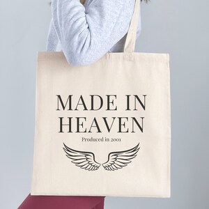 K&ouml;nnte beinhalten: Eine wei&szlig;e Einkaufstasche mit schwarzem Text, der "MADE IN HEAVEN Produced in 2001" lautet, und einem Paar schwarzer Engelsfl&uuml;gel.