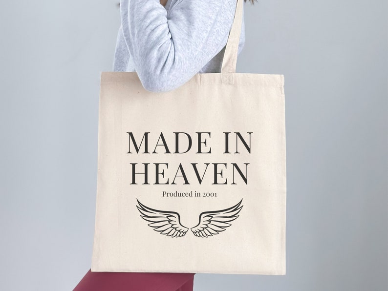 K&ouml;nnte beinhalten: Eine wei&szlig;e Einkaufstasche mit schwarzem Text, der "MADE IN HEAVEN Produced in 2001" lautet, und einem Paar schwarzer Engelsfl&uuml;gel.