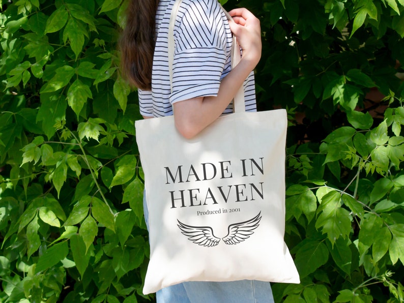 K&ouml;nnte beinhalten: Eine wei&szlig;e Einkaufstasche mit schwarzem Text, der "Made in Heaven Produced in 2001" lautet, und einem Paar schwarzer Engelsfl&uuml;gel.