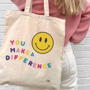 May include: A white canvas tote bag with the text "YOU MAKE A DIFFERENCE" in colorful letters and a yellow smiley face. The bag has a brown leather tag with the letters "EJD" on it.
