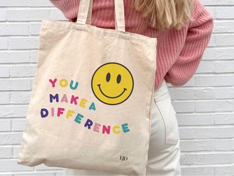 May include: A white canvas tote bag with the text "YOU MAKE A DIFFERENCE" in colorful letters and a yellow smiley face. The bag has a brown leather tag with the letters "EJD" on it.