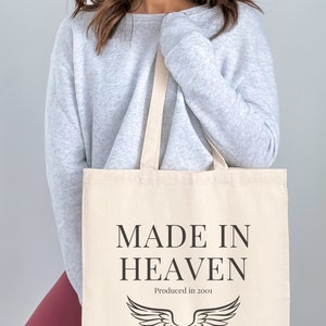 K&ouml;nnte beinhalten: Eine wei&szlig;e Canvas-Tragetasche mit schwarzem Text, der "MADE IN HEAVEN" und "Produced in 2001" lautet. Die Tasche hat ein schwarzes Engelsfl&uuml;gel-Design.