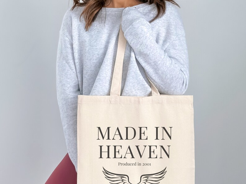 K&ouml;nnte beinhalten: Eine wei&szlig;e Canvas-Tragetasche mit schwarzem Text, der "MADE IN HEAVEN" und "Produced in 2001" lautet. Die Tasche hat ein schwarzes Engelsfl&uuml;gel-Design.