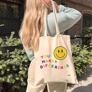May include: A beige canvas tote bag with the text "YOU MAKE A DIFFERENCE" in rainbow colors and a yellow smiley face. The bag has a black logo that says "EJD".