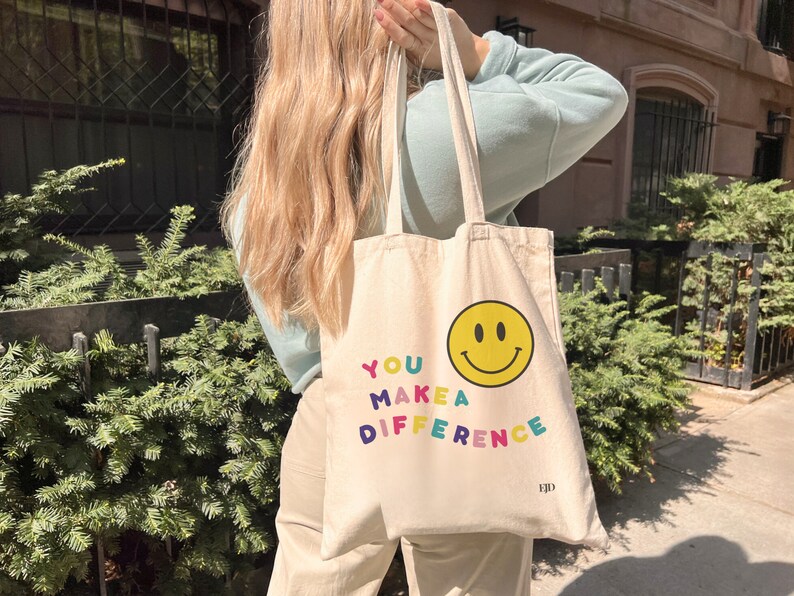 May include: A beige canvas tote bag with the text "YOU MAKE A DIFFERENCE" in rainbow colors and a yellow smiley face. The bag has a black logo that says "EJD".