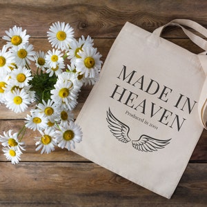 K&ouml;nnte beinhalten: Eine wei&szlig;e Canvas-Tragetasche mit schwarzem Text, der "MADE IN HEAVEN" und "Produced in 2001" lautet. Die Tasche hat schwarze Fl&uuml;gel darauf gedruckt. Ein Strau&szlig; wei&szlig;er G&auml;nsebl&uuml;mchen befindet sich in der oberen linken Ecke des Bildes.