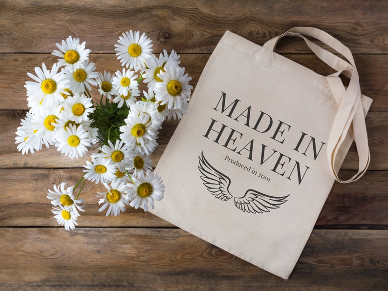 K&ouml;nnte beinhalten: Eine wei&szlig;e Canvas-Tragetasche mit schwarzem Text, der "MADE IN HEAVEN" und "Produced in 2001" lautet. Die Tasche hat schwarze Fl&uuml;gel darauf gedruckt. Ein Strau&szlig; wei&szlig;er G&auml;nsebl&uuml;mchen befindet sich in der oberen linken Ecke des Bildes.