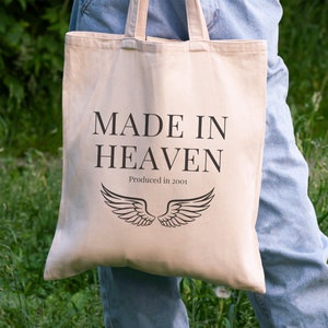 K&ouml;nnte beinhalten: Eine beige Tragetasche mit schwarzem Text, der "MADE IN HEAVEN" und "Produced in 2001" lautet. Die Tasche hat schwarze Fl&uuml;gel darauf gedruckt.