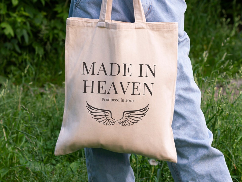 K&ouml;nnte beinhalten: Eine beige Tragetasche mit schwarzem Text, der "MADE IN HEAVEN" und "Produced in 2001" lautet. Die Tasche hat schwarze Fl&uuml;gel darauf gedruckt.