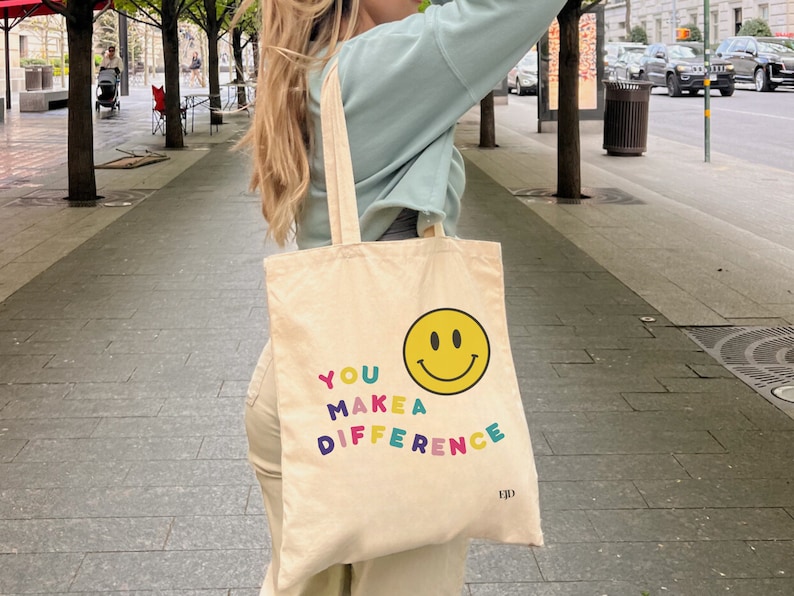 May include: A beige canvas tote bag with a yellow smiley face and the text "YOU MAKE A DIFFERENCE" in colorful letters.