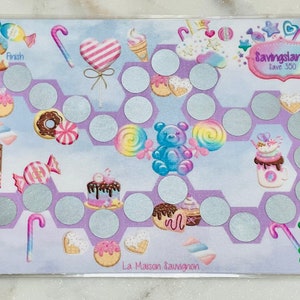 May include: A purple and blue board game with a candy theme. The game board features a gingerbread house, candy canes, lollipops, doughnuts, and bears. The game is called "Savingsland Save 350" and is made by "La Maison Sauvagnon".