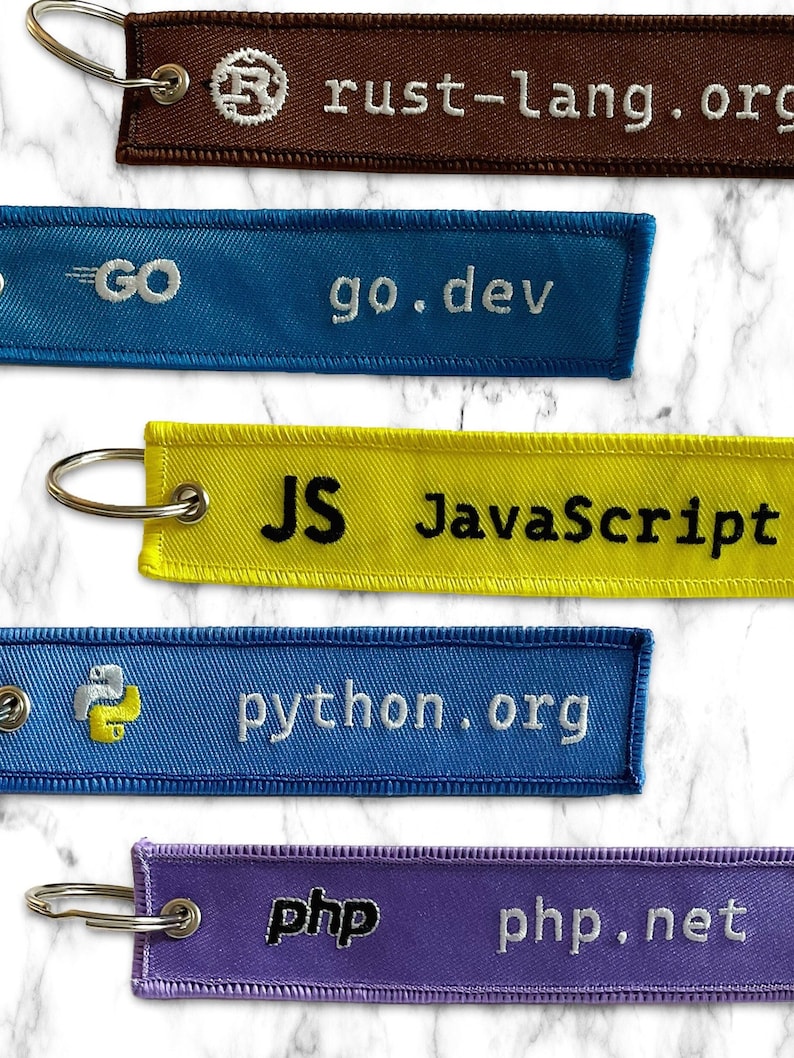 K&ouml;nnte beinhalten: F&uuml;nf bunte Schl&uuml;sselanh&auml;nger mit verschiedenen Programmiersprachen und ihren Websites. Die Schl&uuml;sselanh&auml;nger sind braun, blau, gelb, blau und lila. Der Text auf den Schl&uuml;sselanh&auml;ngern lautet "rust-lang.org", "go.dev", "JS JavaScript", "python.org" und "php php.net".