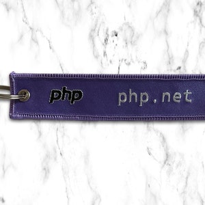 K&ouml;nnte beinhalten: Lilafarbener Stoffschl&uuml;sselanh&auml;nger mit dem Text "php.net" und "php" in schwarzer Schrift.
