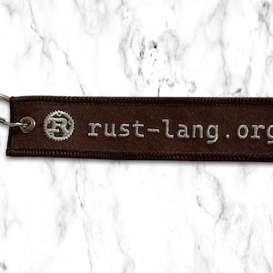 K&ouml;nnte beinhalten: Brauner Stoffschl&uuml;sselanh&auml;nger mit einem silbernen Metallring und dem Text "rust-lang.org" in wei&szlig;er Stickerei.