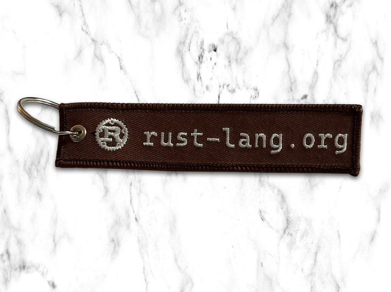 K&ouml;nnte beinhalten: Brauner Stoffschl&uuml;sselanh&auml;nger mit einem silbernen Metallring und dem Text "rust-lang.org" in wei&szlig;er Stickerei.